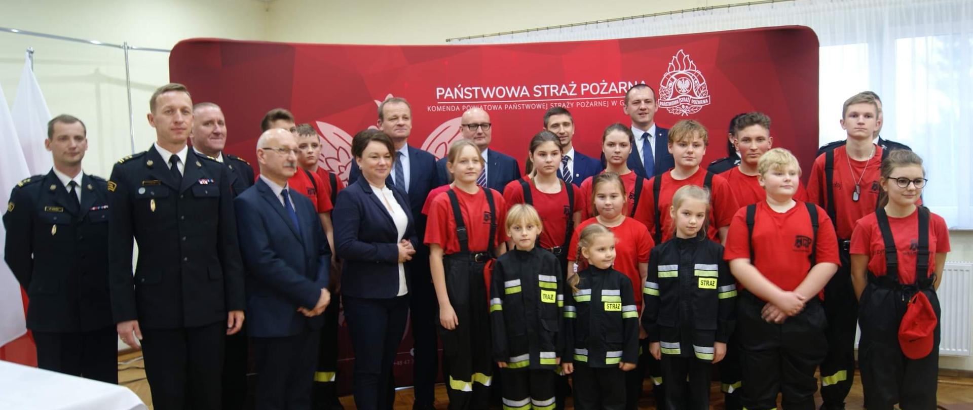 zdjęcie grupowe strażaków i młodzieży w mundurach strażackich , w ostatnim rzędzie kilku mężczyzn w garniturach