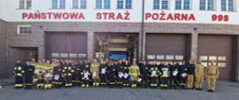 Zdjęcie przedstawia dużą grupę strażaków stojących w szeregu przed budynkiem straży pożarnej w Ostrowi Mazowieckiej, w słoneczny dzień. Budynek jest biały, z ceglanymi elementami i kilkoma dużymi, brązowymi bramami garażowymi. Nad bramami widnieje czerwony napis "PAŃSTWOWA STRAŻ POŻARNA" oraz numer alarmowy "998".
Strażacy, w liczbie około 30, ubrani są w różne typy mundurów: bojowe (czarne z żółtymi i czerwonymi elementami), a także piaskowe mundury polowe. Wielu z nich trzyma w rękach białe teczki lub dyplomy. W centralnej bramie garażowej widoczny jest fragment czarnego samochodu strażackiego. Po lewej stronie zdjęcia widać maszt z flagą Polski. Całość znajduje się na brukowanym placu.
