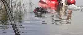 Zdjęcie przedstawia wynurzającego się z jeziora strażaka nurka na tle odpływającej łodzi motorowej Państwowej Straży Pożarnej