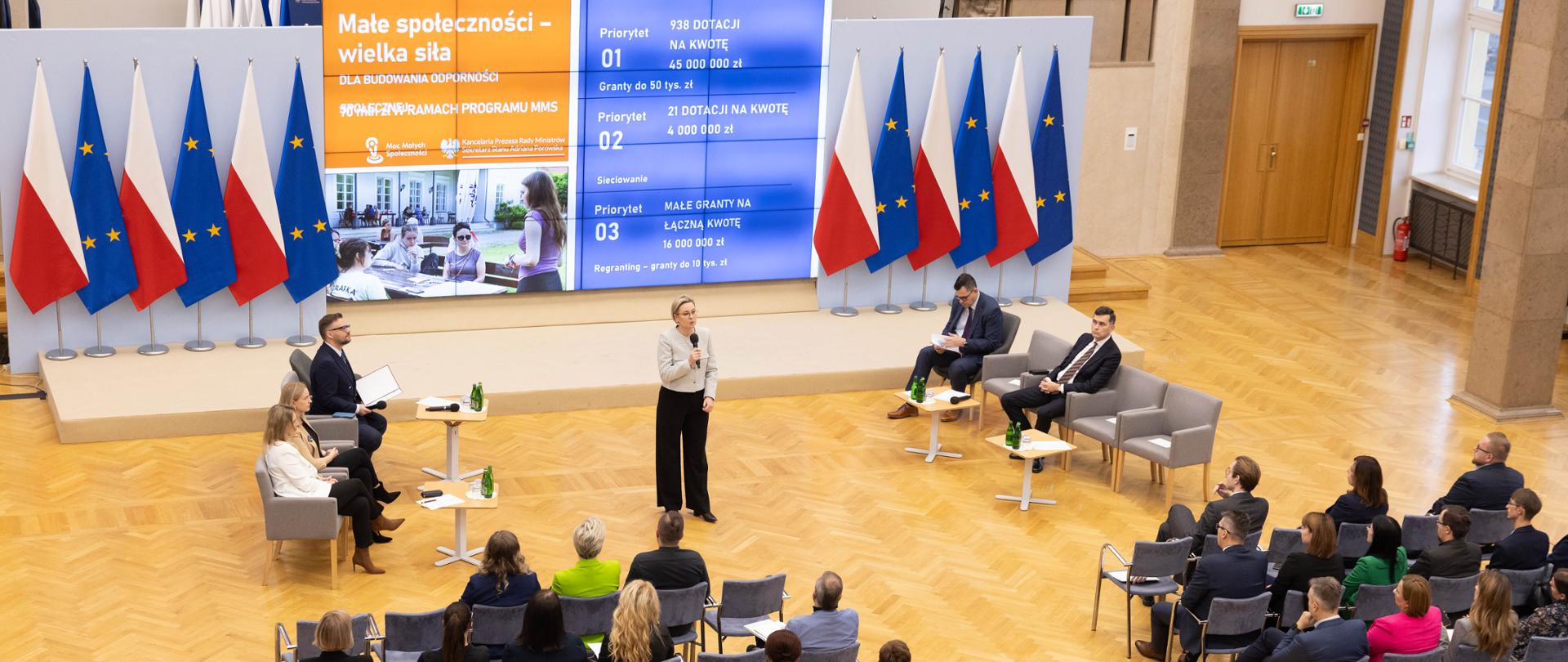 Szeroki kadr, widok sali konferencyjnej. Na zdjęciu Adriana Porowska, Sekretarz Stanu w Kancelarii Prezesa Rady Ministrów, Wiceprzewodnicząca Komitetu ds. Pożytku Publicznego podczas otwarcia konferencji, mówi do uczestników spotkania, którzy zajmują rzędy krzeseł na sali. W tle ekran, na którym wyświetlana jest prezentacja, po obu stronach ekranu flagi Polski i Unii Europejskiej. 