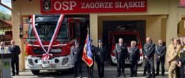 Uroczyste przekazania nowego wozu gaśniczego dla OSP Zagórze Śl. Przy mównicy przemawia Ksiądz. Z prawej strony na baczność stoją strażacy, z czego jeden trzyma sztandar. W tle Wóz strażacki oraz remiza OSP