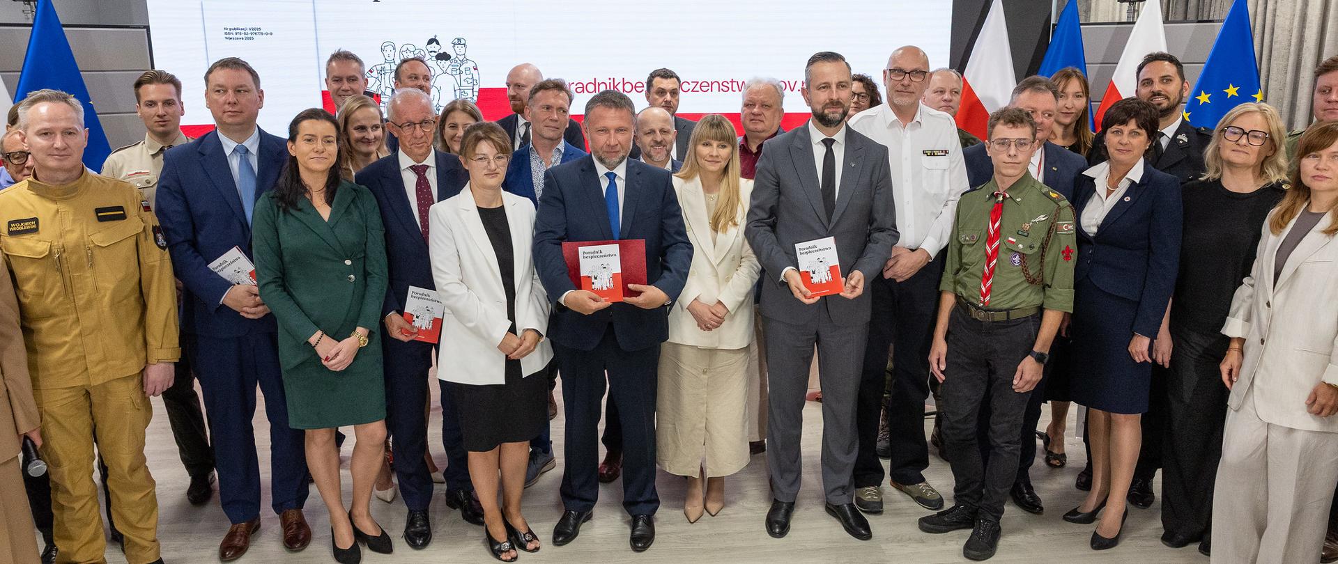 Prezentacja "Poradnika bezpieczeństwa". Na zdjęciu uczestnicy wydarzenia pozują do grupowego zdjęcia. Niektórzy trzymają w dłoniach egzemplarze poradnika. W tle ekran, na którym wyświetlona jest okładka poradnika - biało czerwone tło oraz rysunek, który przedstawia grupę osób. W tle widoczne także flagi Polski i Inii Europejskiej.