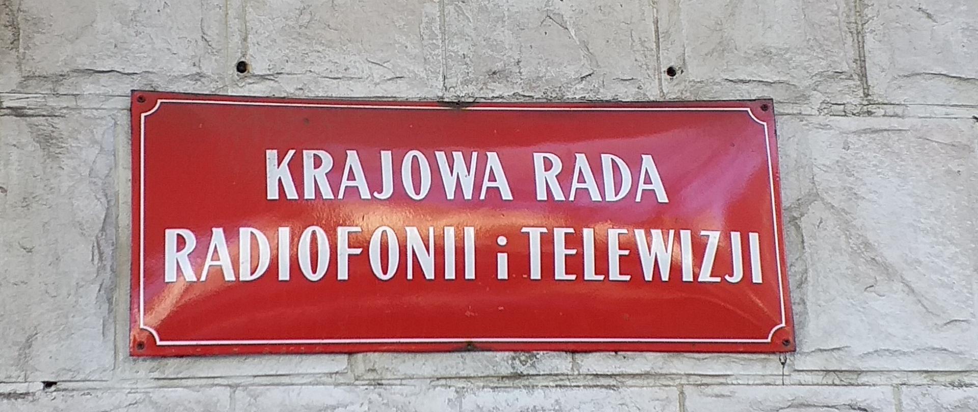 Zdjęcie w poziomie. Podłużna, czerwona tablica z białym napisem "Krajowa Rada Radiofonii i Telewizji"