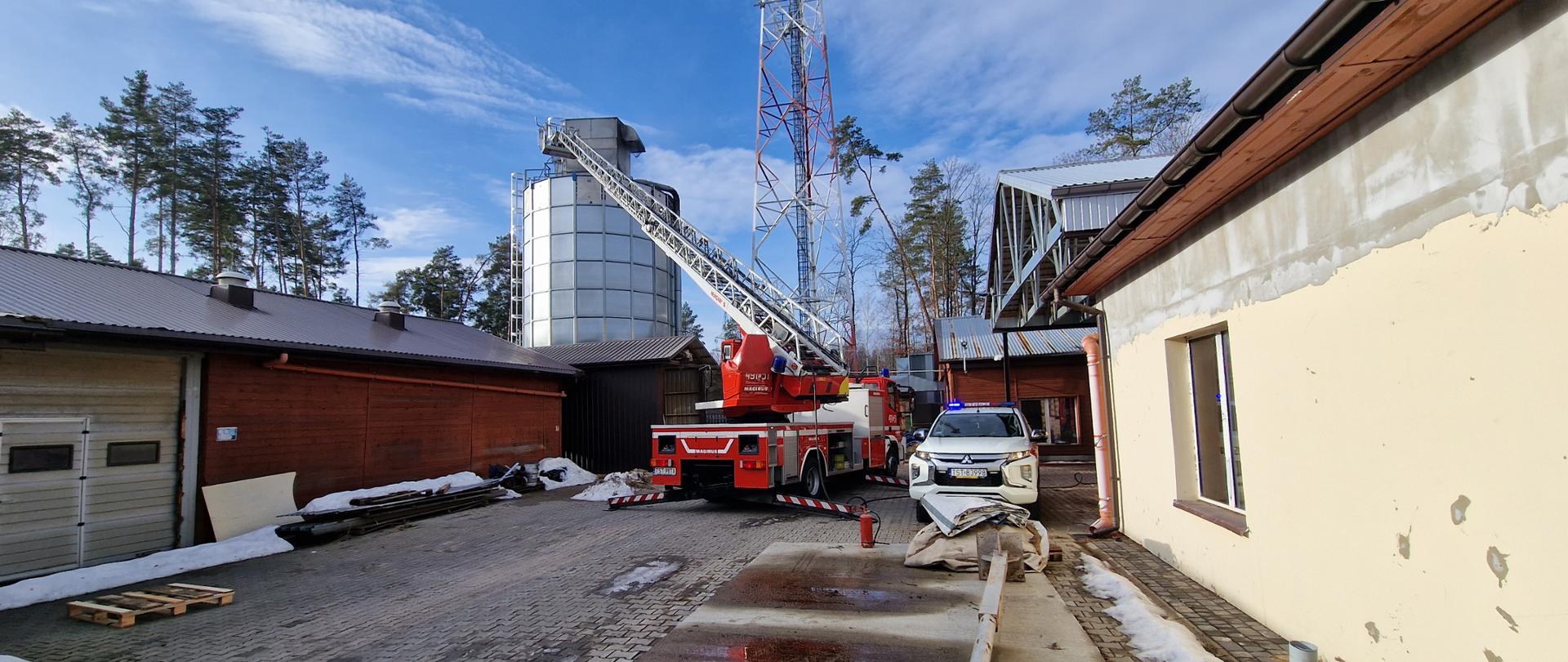 Na pierwszym planie samochody PSP: czerwony wóz z rozłożoną drabiną oraz obok stojący biały samochód ratowniczo-rozpoznawczy, w tle silos z trocinami