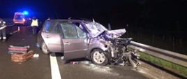 Na zdjęciu widać uszkodzony przód samochodu osobowego, który uderzył w tył naczepy ciężarówki. Z samochodu wyciekły płyny eksploatacyjne. Obok samochodu leżą walizki podróżnicze. Obok pojazdu chodzą policjanci i strażak . W tle widać karetkę pogotowia z włączonymi sygnałami świetlnymi.