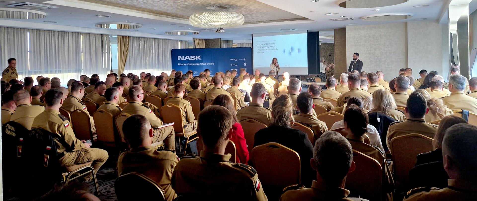 Na zdjęciu widać dużą salę konferencyjną wypełnioną funkcjonariuszami Państwowej Straży Pożarnej. Wszyscy siedzą na krzesłach, zwróceni przodem do mównicy i ekranu projekcyjnego. Mundury strażaków są piaskowe, służbowe, z naszywkami flagi Polski na ramieniu. Na ekranie wyświetlony jest slajd z tytułem prezentacji, a obok ekranu stoi kobieta prowadząca szkolenie oraz mężczyzna,współprowadzący. Za nimi znajduje się niebieskie tło z logo NASK i napisem „Dbamy o bezpieczeństwo w sieci”. Sala jest dobrze oświetlona światłem dziennym wpadającym przez duże okna z zasłonami. Zdjęcie przedstawia moment szkolenia z zakresu cyberbezpieczeństwa dla strażaków.