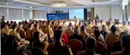 Na zdjęciu widać dużą salę konferencyjną wypełnioną funkcjonariuszami Państwowej Straży Pożarnej. Wszyscy siedzą na krzesłach, zwróceni przodem do mównicy i ekranu projekcyjnego. Mundury strażaków są piaskowe, służbowe, z naszywkami flagi Polski na ramieniu. Na ekranie wyświetlony jest slajd z tytułem prezentacji, a obok ekranu stoi kobieta prowadząca szkolenie oraz mężczyzna,współprowadzący. Za nimi znajduje się niebieskie tło z logo NASK i napisem „Dbamy o bezpieczeństwo w sieci”. Sala jest dobrze oświetlona światłem dziennym wpadającym przez duże okna z zasłonami. Zdjęcie przedstawia moment szkolenia z zakresu cyberbezpieczeństwa dla strażaków.