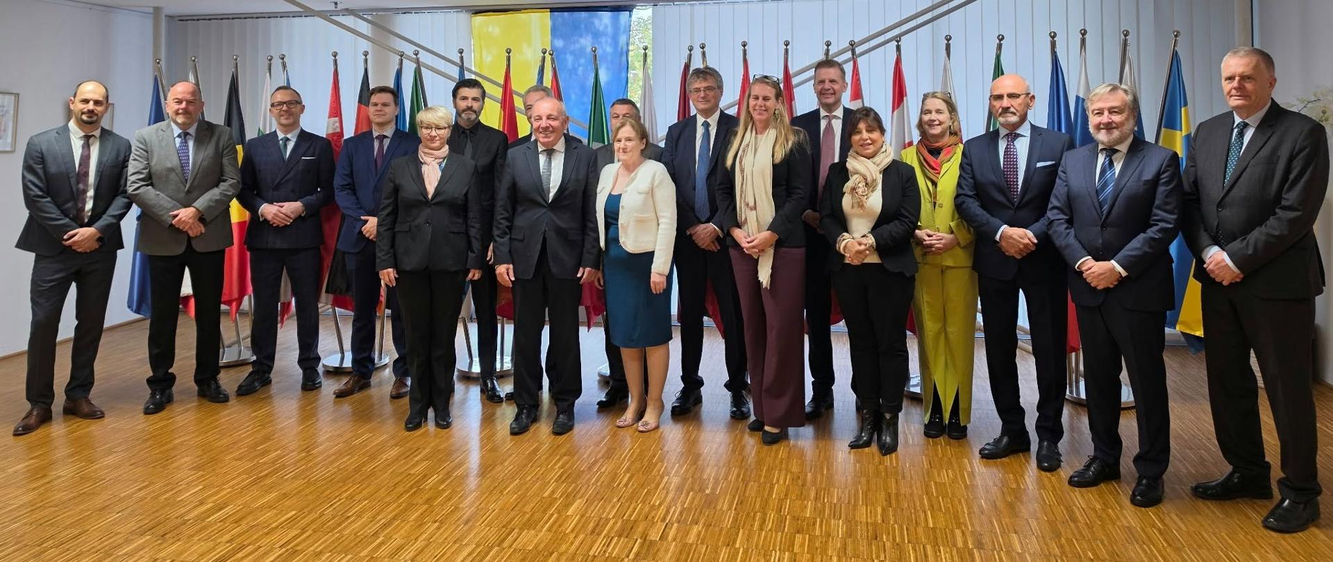 Skupinska fotografija udeležencev diplomatskega srečanja. Petnajst oseb v formalnih oblačilih stoji v vrsti v konferenčni dvorani, pred zastavami evropskih držav. V ospredju je lesen pod, nad njimi pa svetel strop s panelno razsvetljavo.