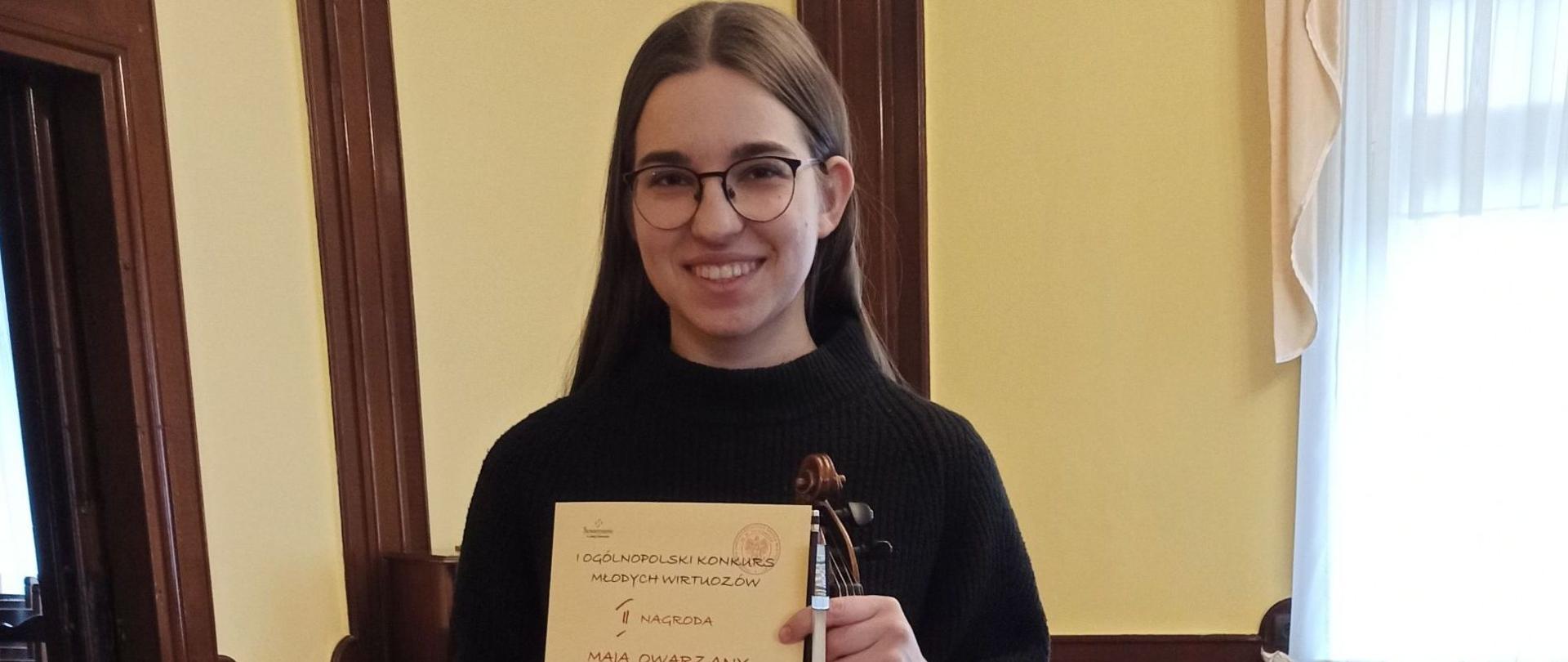 Zdjęcie kolorowe. Dziewczyna w okularach i czarnym golfie trzymająca w dłoniach dyplom oraz skrzypce ze smyczkiem. Dyplom z napisem "I OGÓLNOPOLSKI KONKURS MŁODYCH WIRTUOZÓW II NAGRODA MAJA OWARZANY". W tle wnętrze z drewnianymi panelami na ścianach, wzorzystym dywanem i jasną zasłoną w oknie.
