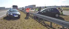 Zderzenie pojazdów na S5. Odcinek ekspresowej "piątki" pomiędzy węzłami Bojanowo i Rawicz. Na pasie zieleni - prostopadle do osi jezdni stoi ciemny samochód osobowy, który przodem uderzył w barierę. Wokół samochodu strażacy i policjanci uczestniczący w akcji. Z lewej strony na pasie zieleni znajduje się nieoznakowany radiowóz policyjny i wozy straży pożarnej. W tle widoczne zabudowania i las.