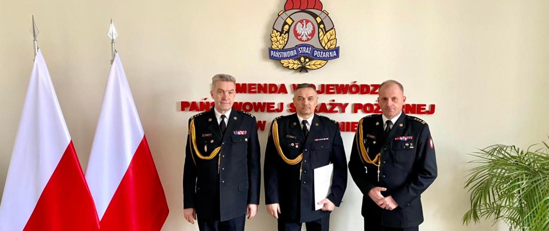 Na zdjęciu trzech mężczyzn, od lewej: Zastępca Zachodniopomorskiego Komendanta Wojewódzkiego PSP st. bryg. Kazimierz Lesisz, Naczelnik Wydziału Organizacji i Nadzoru bryg. Andrzej Dębicki, Zachodniopomorski Komendant Wojewódzki PSP st. bryg. Jarosław Tomczyk
Z lewej strony widoczne flagi, w tle widoczne logo PSP (na ścianie pamiątkowej) z prawej strony widoczny delikatnie kwiat.