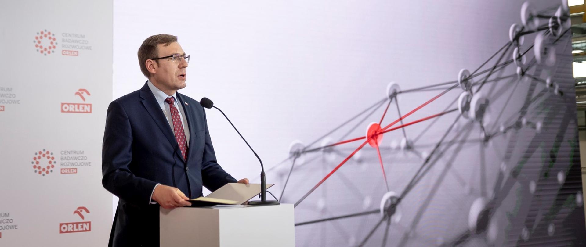 Wiceminister Maciej Małecki podczas uroczystości przemawia z mównicy. W tle ścianka z logo PKN Orlen