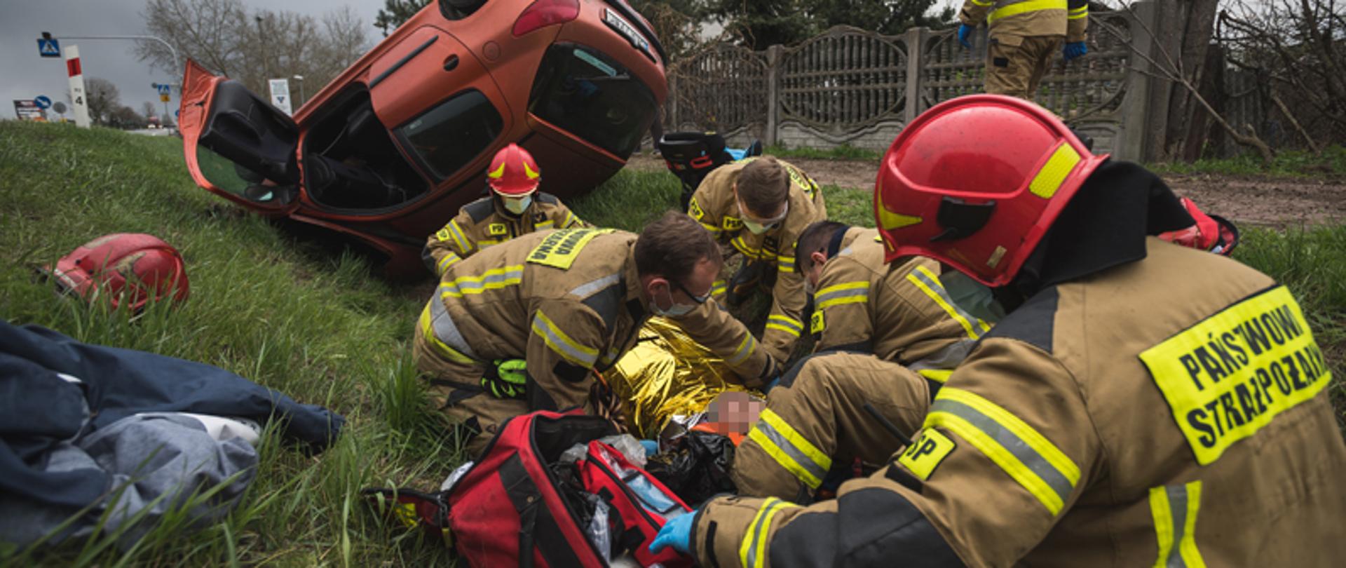 Strażacy udzielają pomocy medycznej poszkodowanej pasażerce, leżącej w przydrożnym rowie. Na drugim planie samochód osobowy leżący na dachu