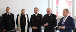 Komendant Powiatowy PSP w Nidzicy wita uczestników spotkania opłatkowego. Obok stoi zastępca komendanta, starosta, ksiądz i strażak.