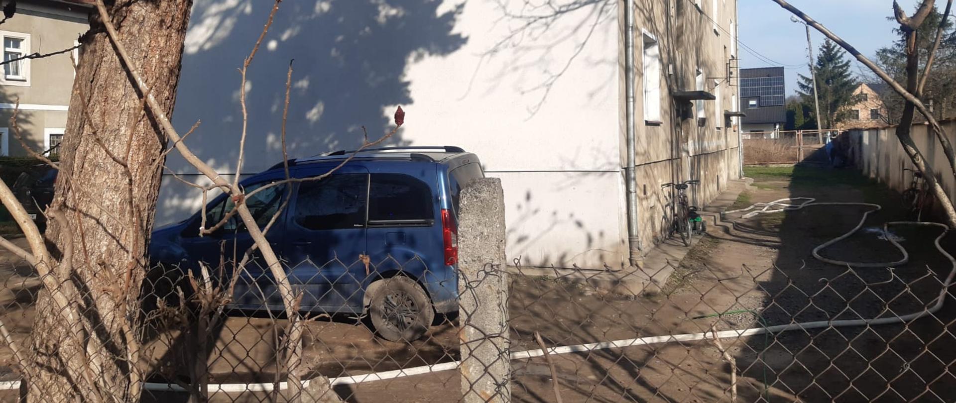 Zdjęcie przedstawia budynek wielorodzinny podczas interwencji straży pożarnej w związku z pożarem jednego z mieszkań. Przed budynkiem stoi zaparkowany niebieski samochód osobowy. Na pierwszym planie widoczne ogrodzenie z siatki. 