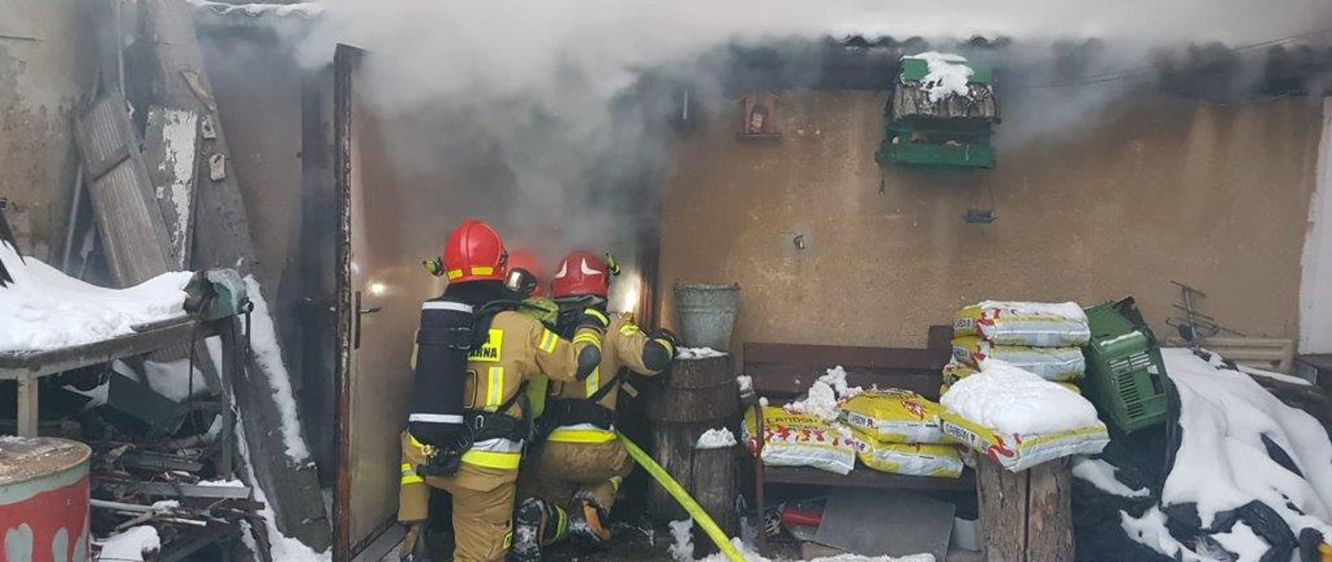 Na zdjęciu widzimy trzech strażaków w pozycji klęczącej, znajdujących się przed wejściem do pomieszczenia kotłowni. Strażacy skierowani tyłem do kadru trzymają wąż strażacki oraz prądownice gaszą pożar. Strażacy mają na sobie ubrania specjalne koloru piaskowego, na głowie czerwone hełmy. Na plecach butle z powietrzem od aparatu ochrony dróg oddechowych koloru czarnego. Z pomieszczenia wydobywa się biały dym. Przed budynkiem widzimy także poukładane worki foliowe. Aura zimowa na ziemi i dachu zalega śnieg. Z lewej strony kadru widzimy część beczki koloru czerwonego.