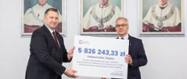 Minister Czarnek i mężczyzna w garniturze stoją i trzymają razem symboliczny czek z napisem 5 826 243,33 zł, za nimi na ścianie wiszą 3 portrety.