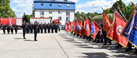 Na zdjęciu widoczna jest uroczystą zbiórka strażaków. Z lewej strony druhowie Ochotniczych Straży Pożarnych prezentują swoje sztandary. Przed sztandarami, w pozycji zasadniczej stoi dwóch funkcjonariuszy PSP, którzy salutują sztandarom OSP. W tle stoją pododdziały strażaków w umundurowaniu galowym, za nimi widoczne są wozy bojowe oraz budynek z niebieskim dachem. Całość odbywa się na otwartym placu przy pięknej, słonecznej pogodzie.
