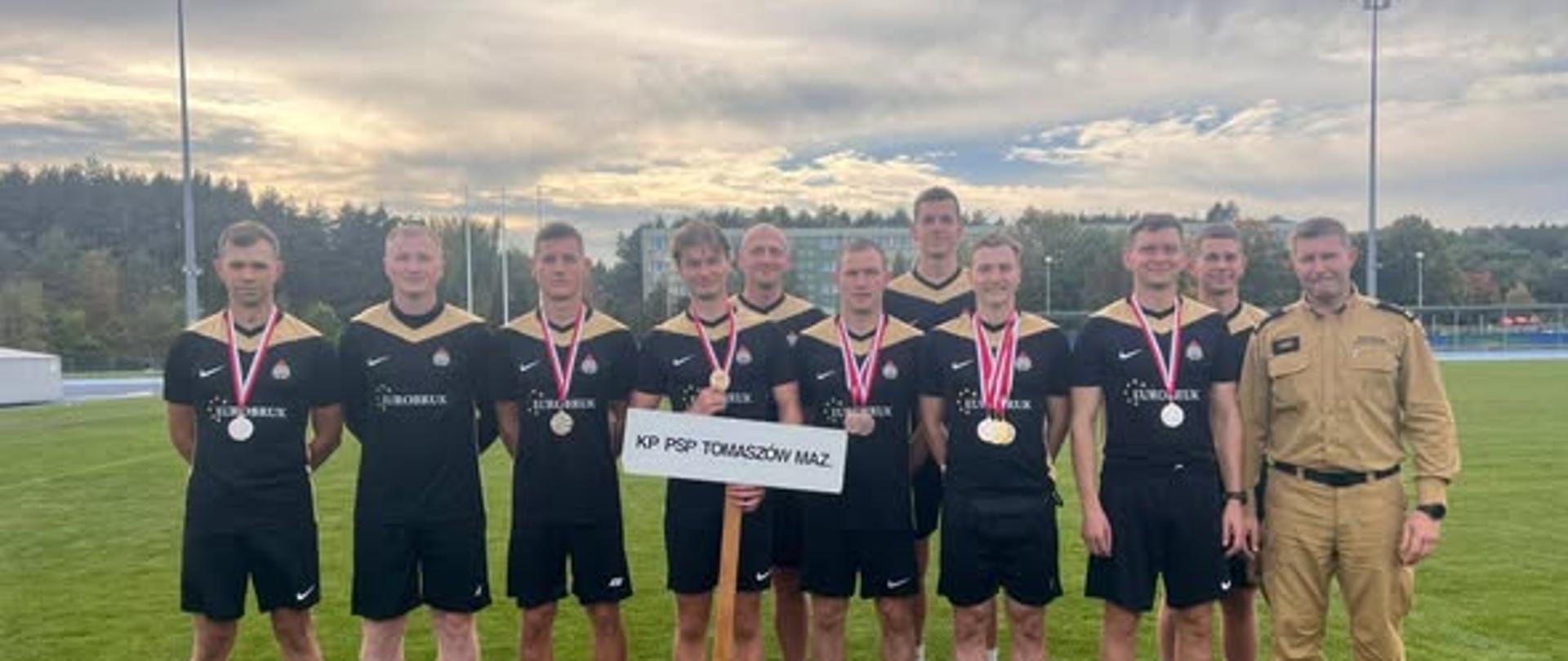 Strażacy stoją na zielonej trawie ubrani w czarno złote stroje. Na szyi mają zawieszone medale. Po prawej stronie stoi mężczyzna ubrany w piaskowy mundur. 