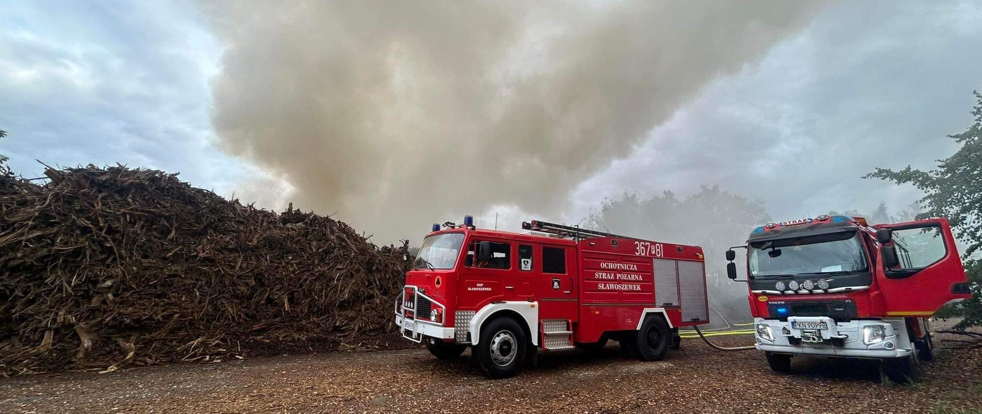 Zdjęcie przedstawia pożar sterty biomasy. Nad biomasą unosi się gęsty dym. Przed stertą biomasy widać dwa samochody strażackie. 