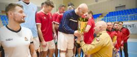 Sportowe podium wypełnione przez trzy drużyny - kilkudziesięciu zawodników w strojach sportowych. Strażak wręcza puchar jednemu z nich, stojącemu na środku. Ten pochyla się w stronę wręczającego. W dali widać krzesła trybuny sportowej. 