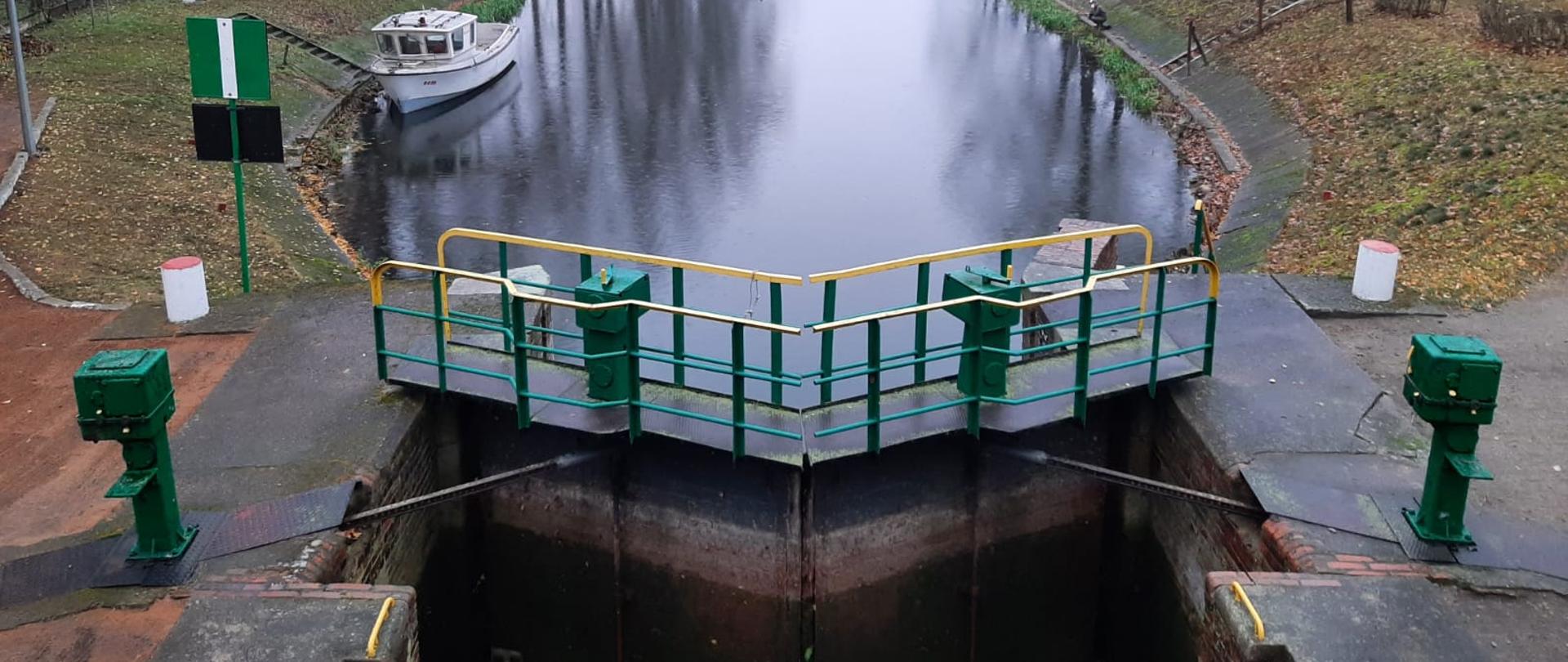 Śluza w Pakości - pierwsza śluza na Górnonoteckim odcinku drogi wodnej Warta – Kanał Bydgoski. Widok z góry na awanport. Na drugim planie, po lewej stronie biały statek o nazwie Brda służący pracownikom Wód Polskich do prac utrzymaniowych. Po prawej stronie siedzący wędkarz nad rzeką Notecią. Po obu brzegach rzeki bezlistne drzewa. W oddali most umiejscowiony nad Notecią. 