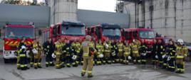 Widok z przodu. Zdjęcie grupowe uczestników ćwiczeń na tle samochodów pożarniczych i zapory wodnej. Tyłem stoi instruktor z PSP.