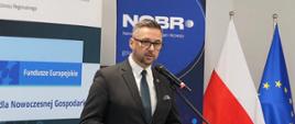 Minister Kulasek stoi i mówi do mikrofonu na stojaku, w ręku trzyma niebieską książeczkę, za nim pionowe banery z napisem NCBR, z prawej pod ścianą flagi Polski i UE.