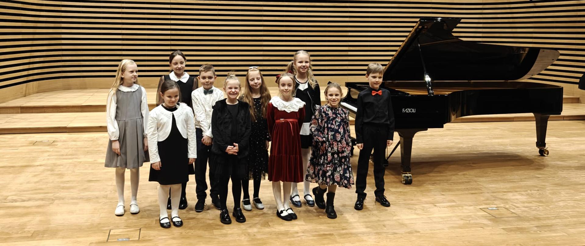 Wszyscy wykonawcy stoją na scenie przy fortepianie.