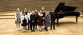 Wszyscy wykonawcy stoją na scenie przy fortepianie.