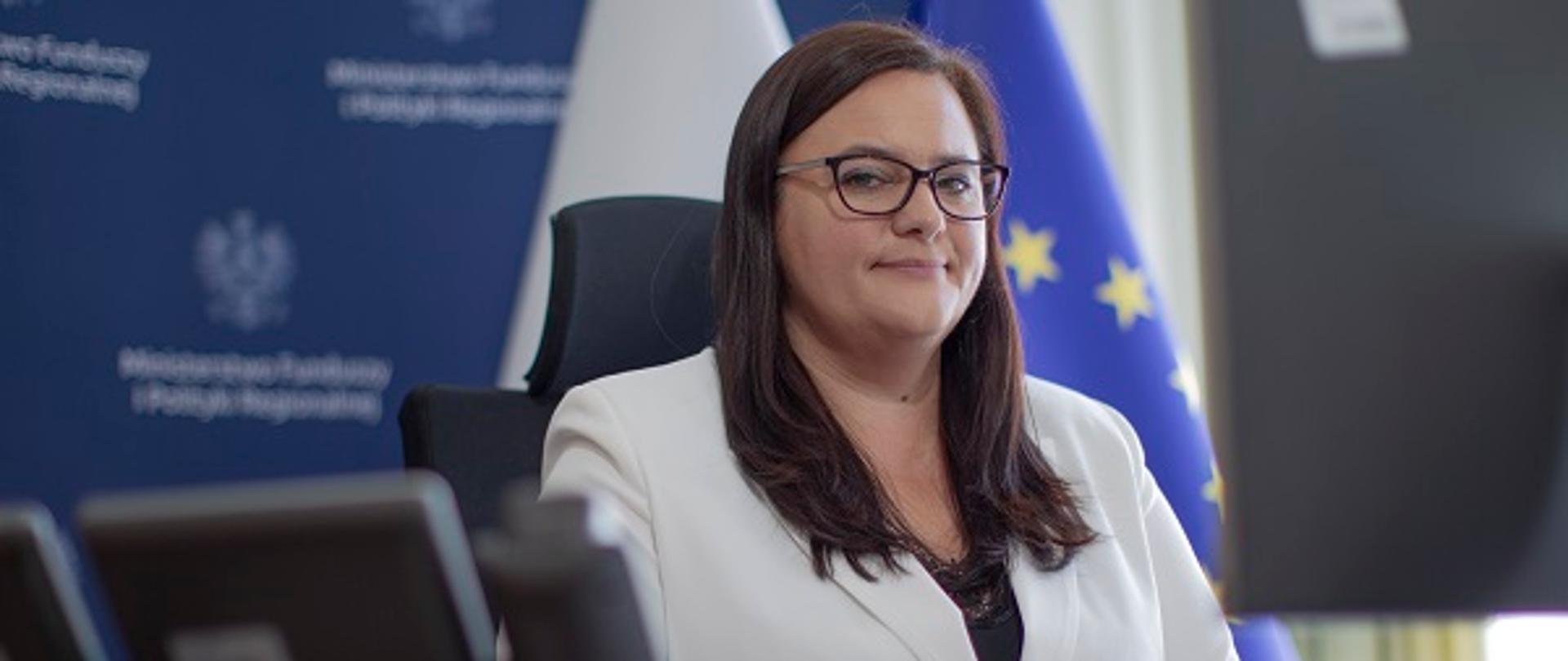Minister Małgorzata Jarosińska-Jedynak siedzi siedzi przy biurku przed laptopem. Z tyłu flagi Polski i UE.