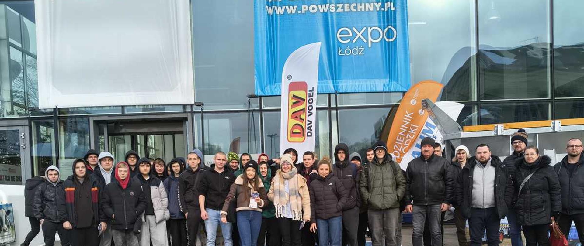 Na zdjęciu znajduje sie grupa ok 30 osób na tle hali EXPO ŁÓdź. Wszyscy ubrani w ciuchy zimowe - kurtki, czapki, kaptury. Stoją na miejscu parkinfowym dla niepełnosprawnych - niebieska koperta ze znaczkiem inwaliczkim. W tle znajduje sie hala z napisem "EXPO ŁÓDŹ"