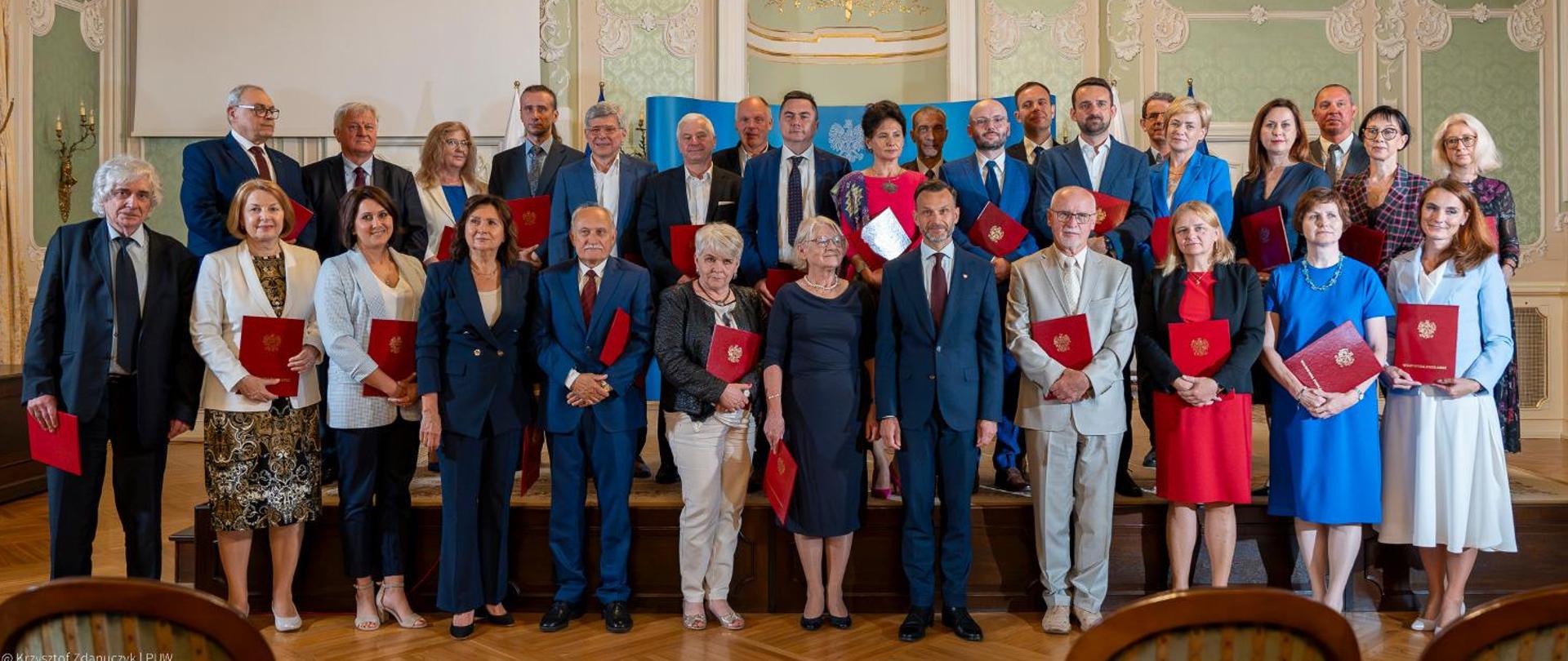 Elegancko ubrani ludzie stoją w dwóch rzędach pod kryształowym żyrandolem w reprezentacyjnej sali Pałacu Branickich w Białymstoku. W środku wojewoda podlaski Jacek Brzozowski. Pozostali trzymają w rękach czerwone teczki ze złotym orłem na okładkach.