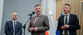 Minister Wieczorek stoi i mówi do mikrofonu, obok niego stoi wiceminister Gdula i mężczyzna w garniturze w granatową kratę.
