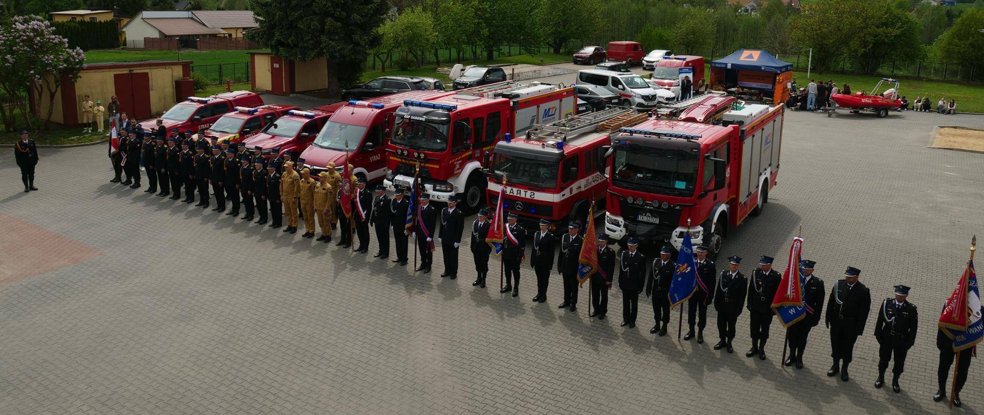 Pododdział strażaków, na tle wozów strażackich. Widok z góry. Jest to jakaś uroczystość strażacka. Strażacy w mundurach wyjściowych ze sztandarami 