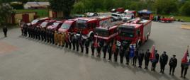 Pododdział strażaków, na tle wozów strażackich. Widok z góry. Jest to jakaś uroczystość strażacka. Strażacy w mundurach wyjściowych ze sztandarami 