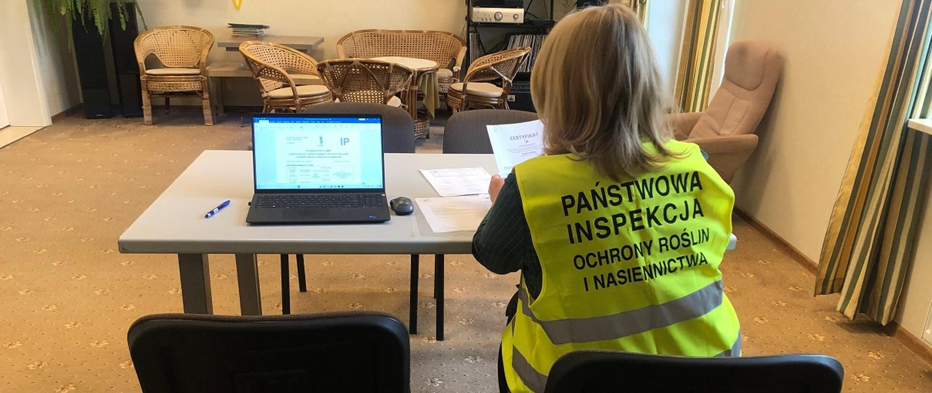 Inspektor WIORiN kontroluje dokumenty w jednostce certyfikującej. Siedzi przy stole w kamizelce odblaskowej z logo PIORiN, trzyma w ręku certyfikat, obok leży laptop z otwartym protokołem kontroli