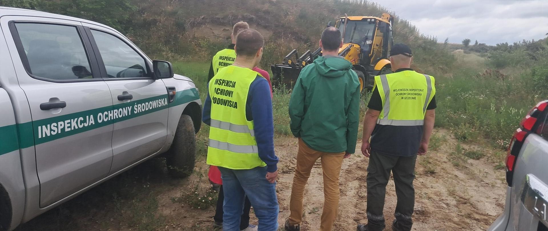 Inspektorzy IOŚ z przedstawicielami gminy przed przystąpieniem do czynności na kontrolowanym terenie. Widoczne pojazdy służbowe oraz koparka. 