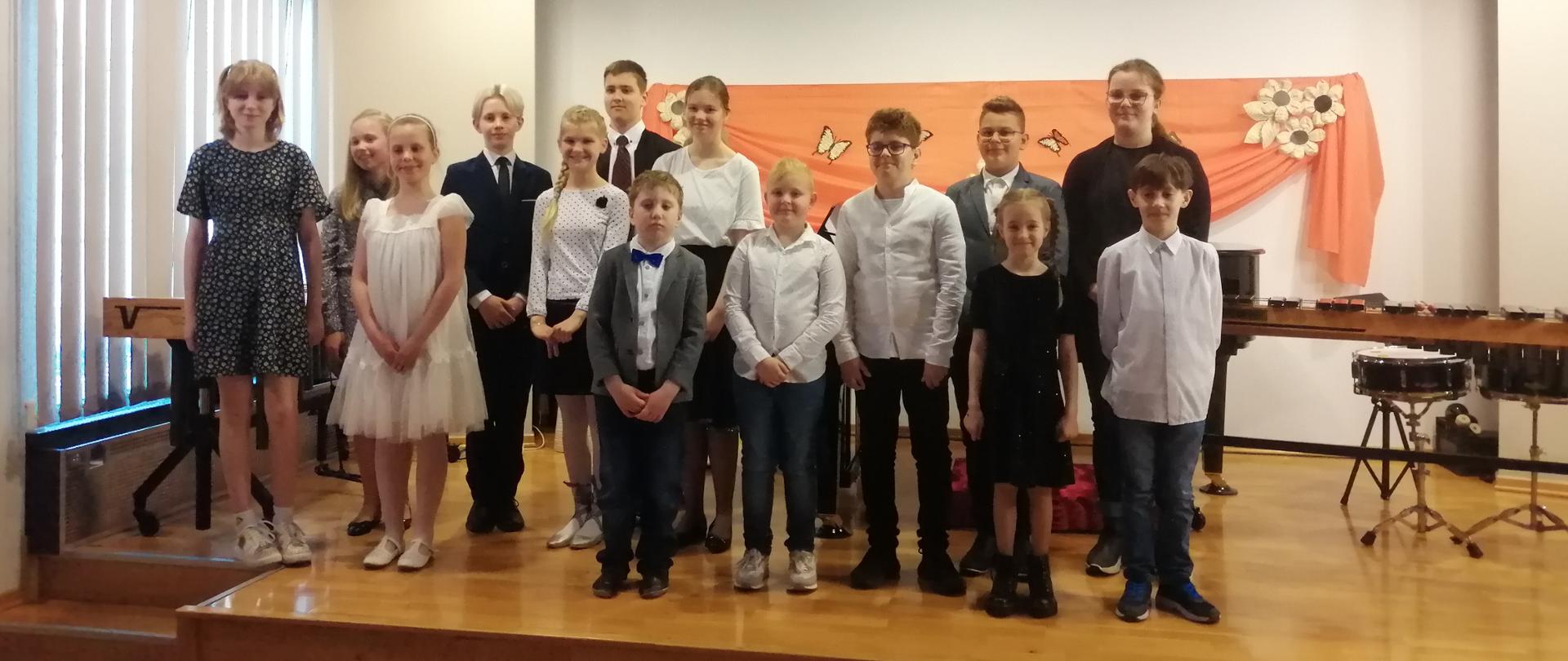 Zdjęcie wykonanie na scenie. Przedstawia ono uczniów szkoły, którzy wystąpili podczas koncert. W tle dostrzegamy instrumenty perkusyjne oaz dekorację wykonaną z pomarańczowego materiału, na którym umocowano kwiaty i motyle.