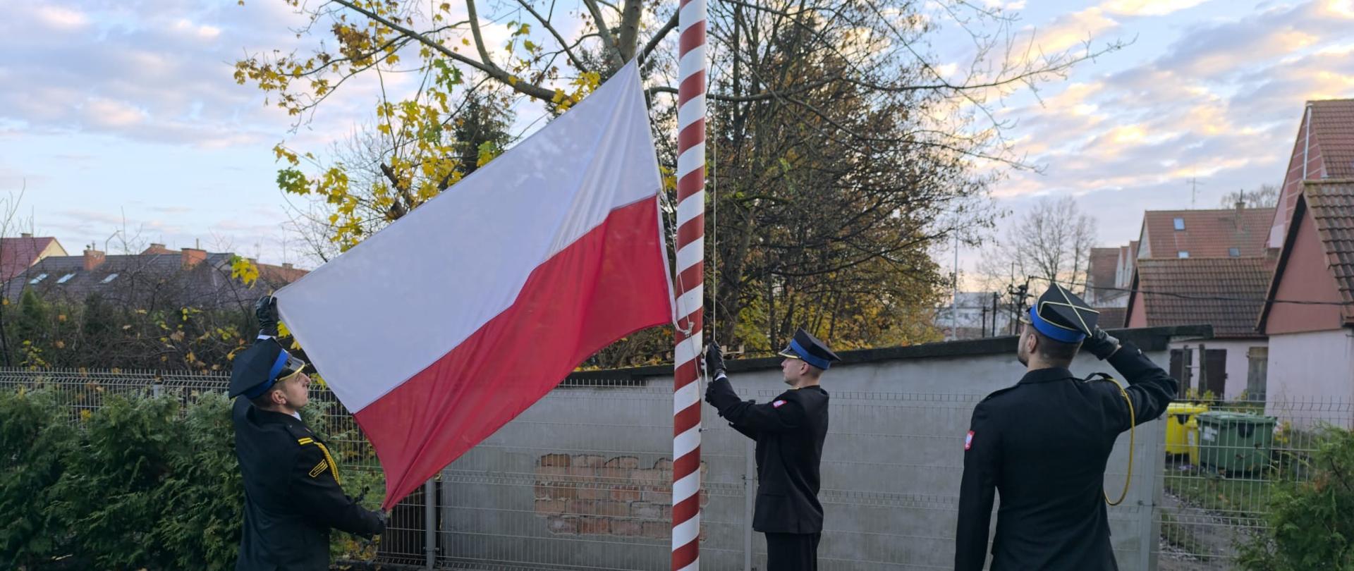 Poczet flagowy uroczyście montuje flagę państwową do linki nośnej masztu flagowego. Jeden z trzech strażaków wchodzących w skład pocztu flagowego salutuje.