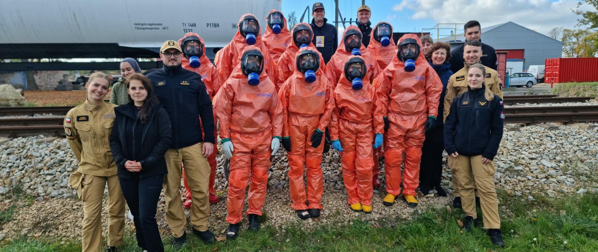 Poligon szkolny, pamiątkowe zdjęcie grupowe strażaków z Ukrainy ubranych w specjalistyczny kombinezon chemiczny (10 osób) w towarzystwie instruktorów oraz tłumaczy