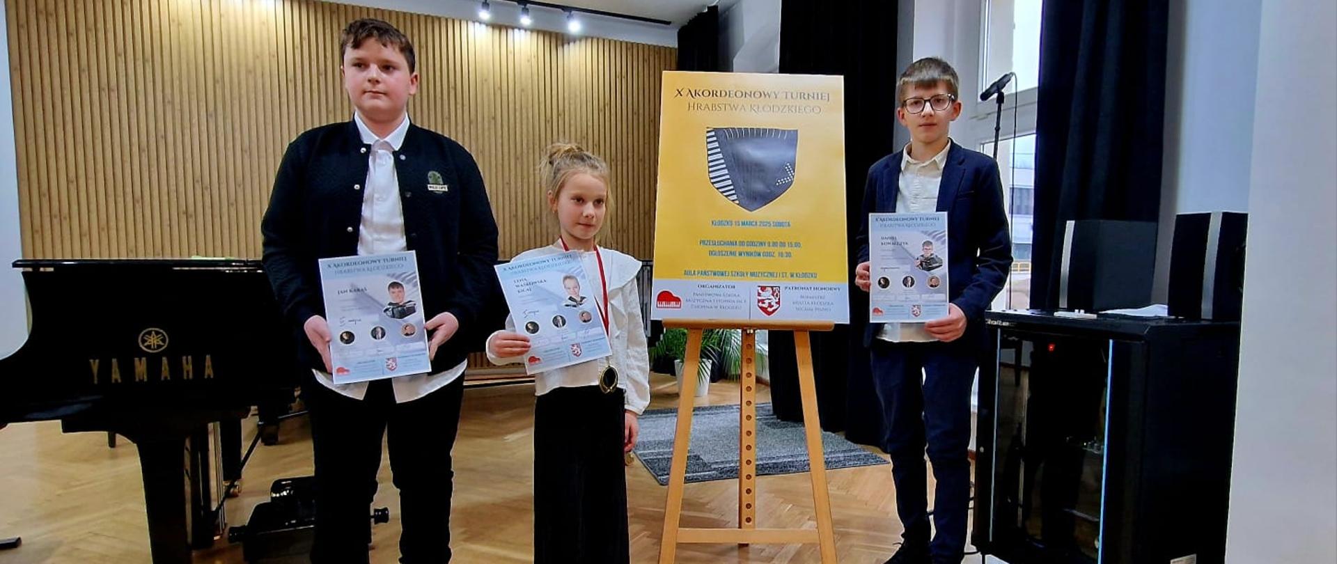 Zdjęcie przedstawia trzech laureatów konkursu akordeonowego trzymających dyplomy na tle plakatu X Akordeonowego Turnieju Hrabstwa Kłodzkiego 2025 i fortepianu