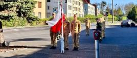 Kilka osób w mundurach stoi w półokręgu na placu przed budynkiem (prawdopodobnie remizą). Część z nich salutuje w kierunku masztu z biało-czerwoną flagą. Na ziemi widoczne są długie cienie, co wskazuje na wczesną porę dnia. W tle znajdują się drzewa i zabudowa osiedlowa.