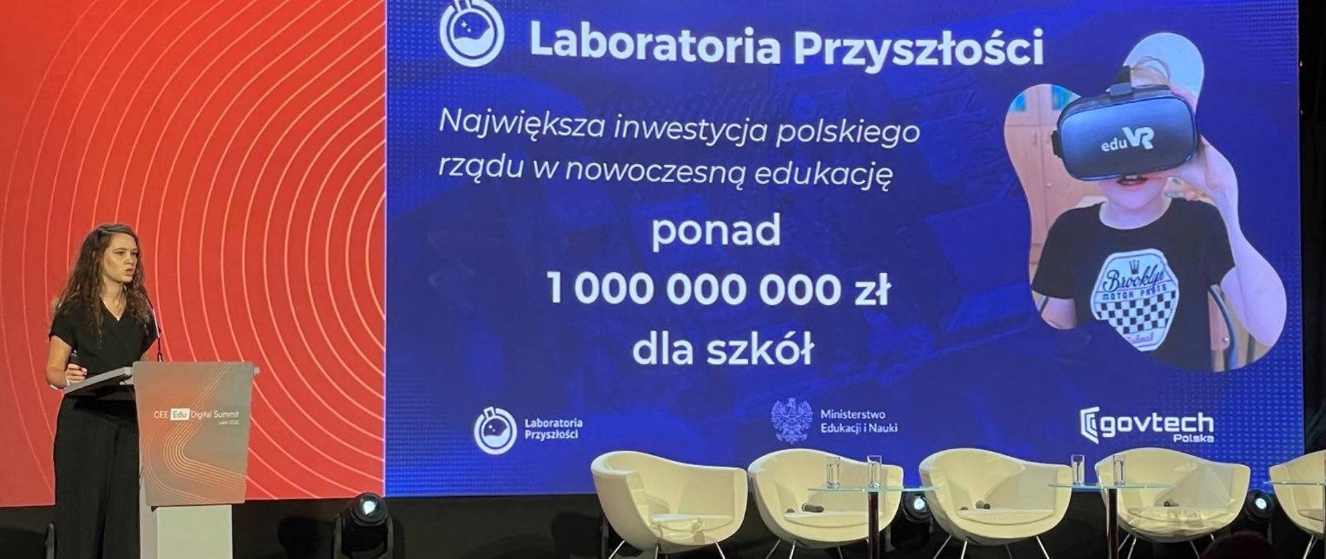 Przed zaciemnioną salą, w której w rzędach siedzą ludzie, stoi ciemno ubrana kobieta i mówi do mikrofonu, za nią kolorowa ściana z napisem Laboratoria przyszłości.