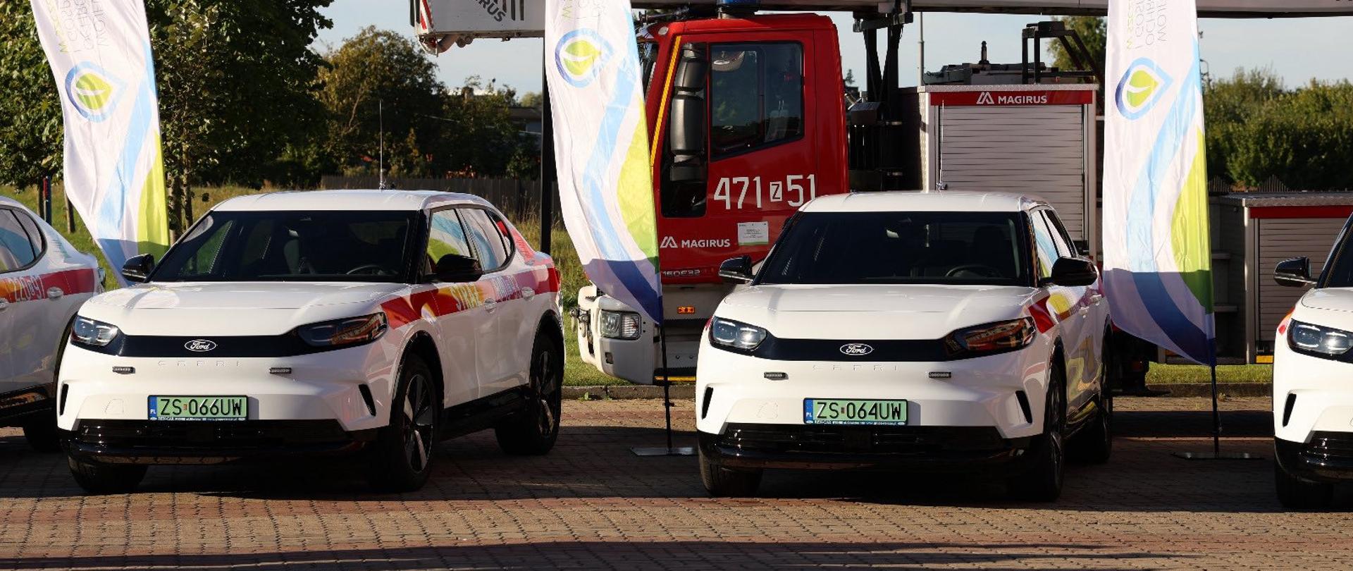 Na zdjęciu widzimy kilka samochodów i wóz strażacki ustawione obok siebie na placu z kostki brukowej. Na pierwszym planie znajdują się dwa białe samochody marki Ford, oba z zielonymi tablicami rejestracyjnymi. Oznacza to, że są to auta elektryczne. Na bokach samochodów widać kolorowe oznaczenia. Za nimi stoi duży czerwony wóz strażacki z drabiną. Po obu stronach samochodów rozstawione są pionowe flagi reklamowe z kolorowymi logotypami i napisami Wojewódzkiego Funduszu Ochrony Środowiska i Gospodarki Wodnej. W tle widać drzewa i niebo w słoneczny dzień.