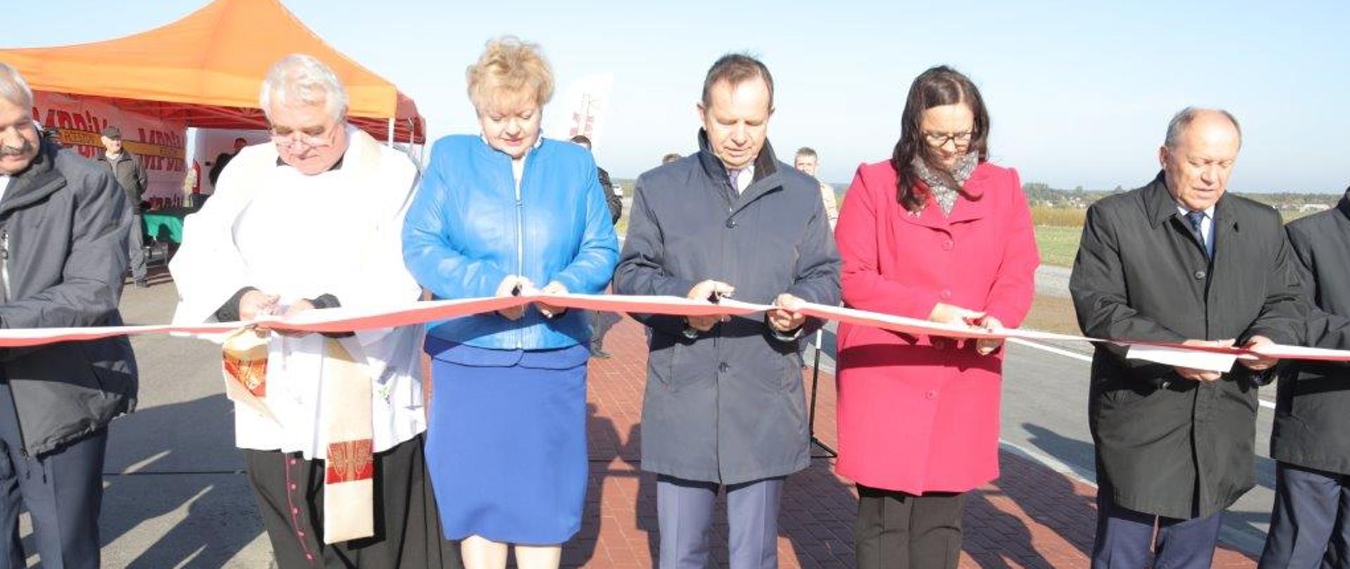 Kilka osób przecina biało-czerwoną wstęgę na chodniku przy obwodnicy; druga z prawej w różowym płaszczu stoi wiceminister Jarosińska-Jedynak, trzeci z prawej marszałek województwa podkarpackiego Władysław Ortyl