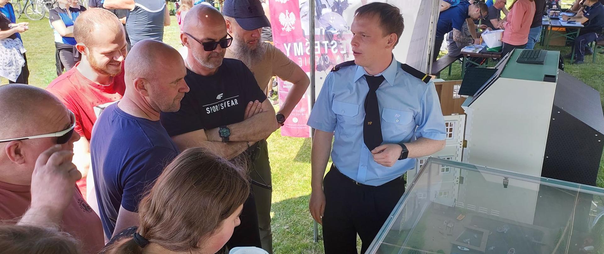 Piknik w Żylicach. Plenerowa remiza strażacka na miejscowym boisku sportowym. Strażak w niebieskiej koszuli prezentuje uczestnikom rajdu rowerowego mobilny symulator zagrożeń i opowiada o zagrożeniach leśnych i wodnych. Jedna z osób trzyma plastikowa miseczkę z zupą. W tle pozostali uczestnicy pikniku oraz winder promujący KP PSP w Rawiczu. 
