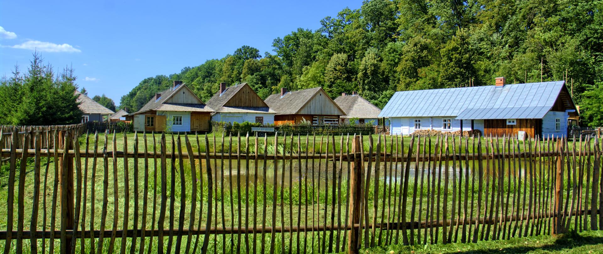 Ilustracja przykładowa Pixabay. Na zdjęciu kilka domków wiejskich, na pierwszym planie drewniany płot. W tle zieleń miejska.