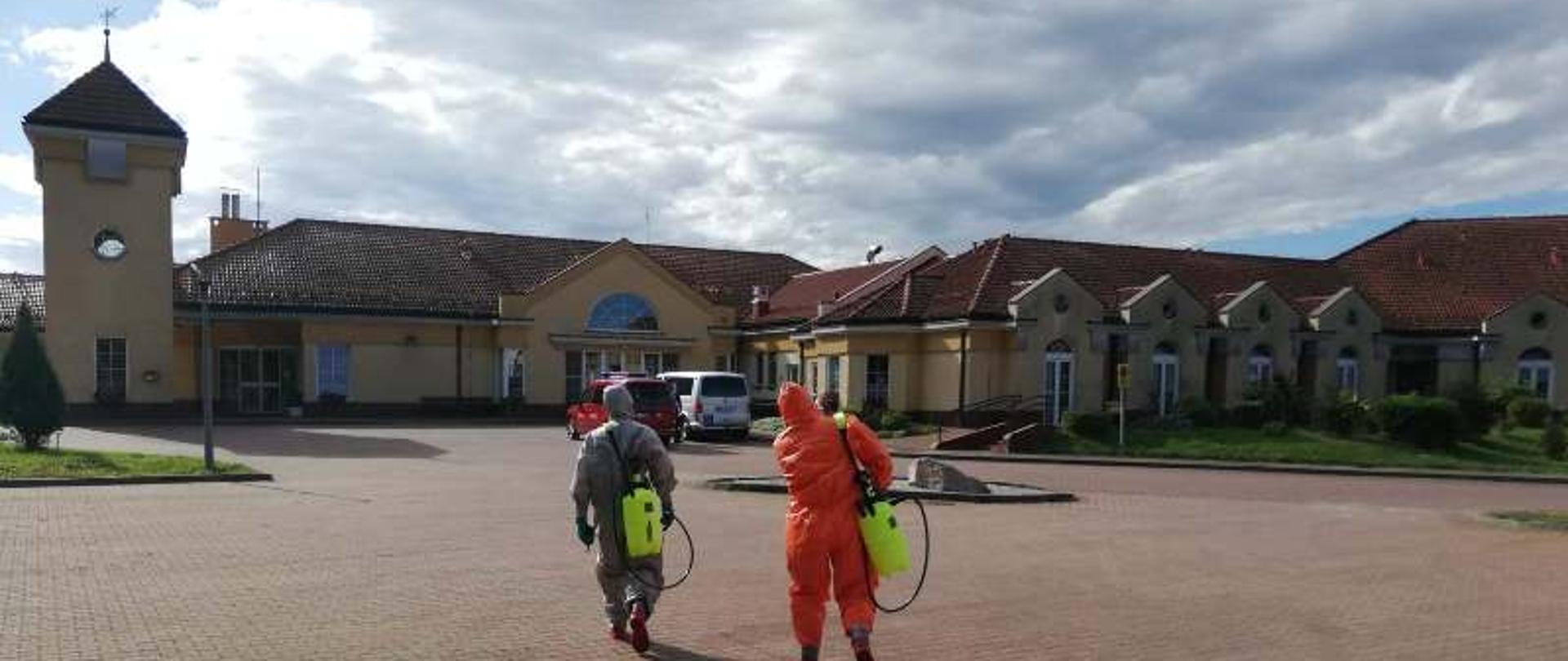 Na pierwszym planie widać dwóch ratowników, którzy zmierzają do siedziby placówki celem podjęcia działań dezynfekcyjnych w obiekcie. Na drugim planie widać obiekt domu pomocy społecznej. Działaniom sprzyja słoneczna pogoda.