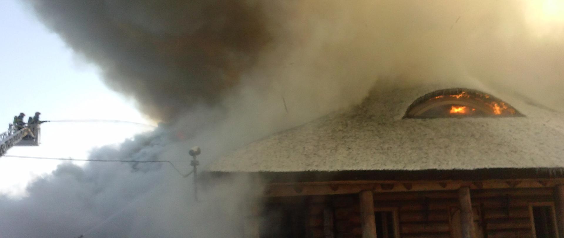 Obraz przedstawia płonący drewniany budynek pokryty strzechą